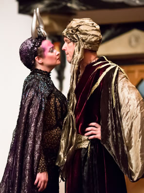 Titania in black glittery gown and silver horns on a 20s black cap stares breast to breast at Oberon in blackand purple robe, gold cape, and gold turban, with his hands on his hips