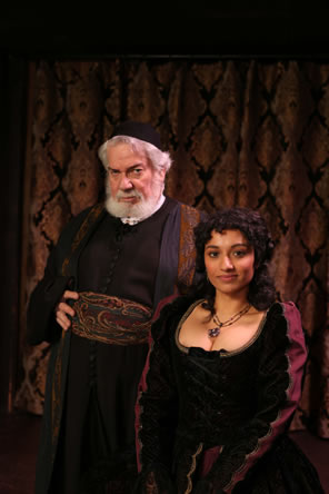 White bearded Shylock in black robe and skullcap with paisley patterned robe and belt poses with Portia wearing a black dress with gold trim and purple sleeves.