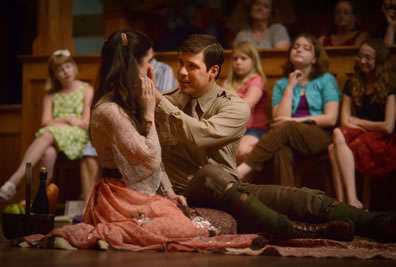 Bertram in army uniform has his hands on Diana's cheek (and her hand is holding his) as they sit on a picnic blanket with members of the audience in the background; she's in a soft orange country dress