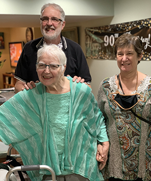 Carol Kelly in a green blouse and house jacket holding hands  with Sarah in a colorful tapestry dress and me in black polo shirt with my hands on Carol's shoulders. In the background is a 90th Birthday! banner