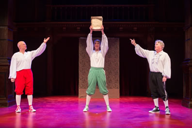 Spencer holds up the play manuscript as Martin and Tichenor flanking him point to it. All are wearing white puffy shirts, knee britches, white stockings, and tennis shoes: Martin in red pants and shoes, Spencer in green pants and shoes, Tichenor in black pants and shoes