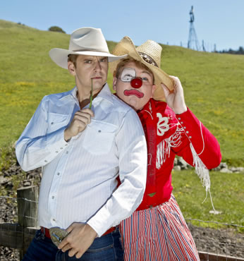 Cowboy in whit shirt, white cowboy hat, jeans and hand gripping huge beltbuckle while the other hand holds a tall grass to his mouth, rodeo clown in red next to him, oil derrick in backgorund