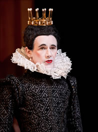 Man wearing woman's makeup in Elizabethan black dress with large ruff colar, gold crown with upright prongs on tight black wig