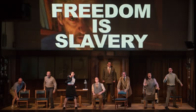 The cast stand and crouch next to chairs, shouting, except Winston sitting, and O'Brien standing at the back in front of the office canteen door; overhead is a video image of the Brotherhood leader with "Freedom Is Slavery" in bold white letters sumperimposed over him