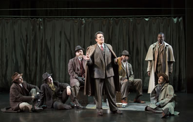 Jaques stands in the center, talking, with two men sitting on the ground and one on a box behind him to his right, another man sitting on a box behind him to his left, the Duke standing in the back and another man sitting on the ground to Jaques left, with the curtains of Arden across the back of the stage, and a blanket spread out on the floor with a coffeepot in the center.