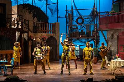 The musicians in various yellow garb stand lined up across the width of the stage from cafe table and chair on the left to a home's tableclothed table and chair to the right. Behind them is the set of Ephesus, with building facades featuring doors and balconys, and a scaffold at the back covered in fishing nets and crates with a boat under construction underneath. The back screen is a twilight blue.