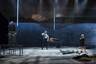 With bodies lying on the stage, the Weird Sisters talk on cell phones, one in combat gear standing on a table to the left, one in white shirt, black pants  and shoulder holster standing at the back center, the woman in business suit and skirt standing to the right.