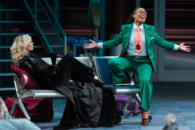 Viola, wearing a green jacket and pants, blue shirt, and bright orange tie with a giant ladybug at the bottom, sits on the arm at one end of a three-seat airport lobby bench, one foot on the ground, one on a seat, arms outspread as she talks to the air while Olivia, stretched out on the other end of the bench and wearing black pant suit and floor-length black coat watches in fascination.