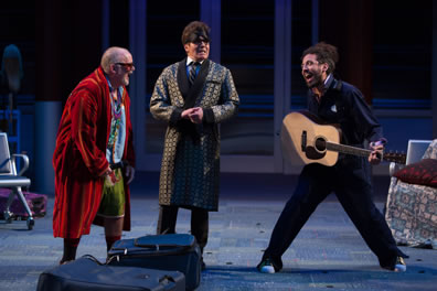 Toby in red robe, blue shurt, greetn shorts and wearing glasses, Malvolio in silk robe over vest, tie, white-colllared blue shirt and gray checked pants and wearing a night eye visor, and Feste, legs splayed as he's holding a guitar, wearing blue utility coveralls; luggage is strewn on the floor.