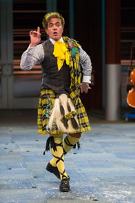 Malvolio steps forward daintly with finger pointed up as he wears a yellow and black plaid quilt, sash and hat, his gray vest and blue striped shirt, plus yellow stockings with cross-garters.