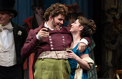 Production photo of Jos hugging Becky, both with drinks in their hands, as tuxedo-wearing stage manager is to the left.