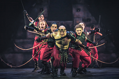 Antony, torso bared and wearing red pants with an amrored belts and black shows, leans forward with ropes tied around his biceps, and angling out behind him are his crew, dressed in black breastplates, red pants, and black boots, all pulling ropes with one hand and holdin swords or poles int heir other hand. Their positions imply the bow of a boat. The pyramid set is in the shadowy background. 