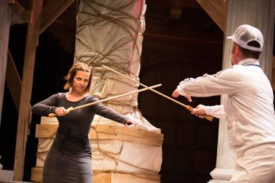 Joan in tight-fitting gray dress and Dauphine in white shirt, slacks, and farmer's cap face off to fence with wooden swords; background is wrapped pillars