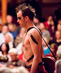 Chris Johnston as Arial in The Tempest with spikey hair, heavy black eye shadow, wearing no shirt, and a mandolin strapped over his should and hanging by his side with the audience in the background.