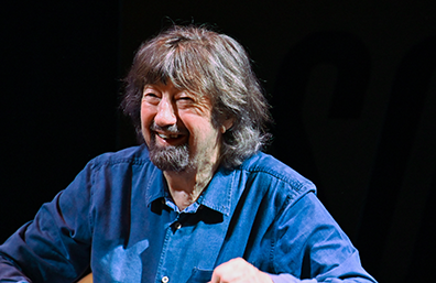 Portrait of Sir trevor Nunn, smiling, wearinga blue denim shirt against a black background